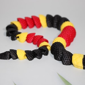 May include: A coiled, stylized snake toy in black, red, and yellow. The segmented body is arranged in a curved pattern, with the head and tail pointing in opposite directions. The snake's head is yellow and black.