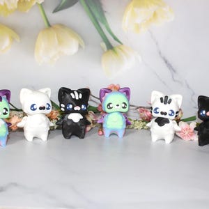 May include: Six colorful cat figurines with big eyes and small arms are arranged in a row. The cats have different color schemes, including white, black, and pastel rainbow hues. The background features flowers and a marble surface.