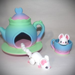 May include: A whimsical, pastel-colored teapot with a heart-shaped lid, a matching teacup, and a white mouse figurine. The teapot has a large opening, and the teacup holds a small mouse. The mouse has pink ears and feet.