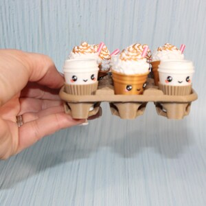 May include: A hand holds a cardboard egg carton containing six miniature coffee cups. Three cups are white with brown sleeves, and three are gold-colored. Each cup has a cute face, whipped cream topping, and a pink straw.