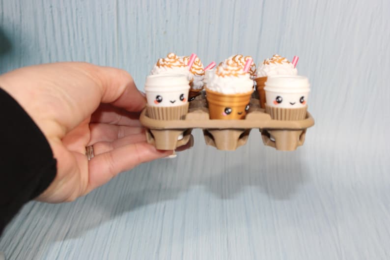 May include: A hand holds a cardboard egg carton containing six miniature coffee cups. Three cups are white with brown sleeves, and three are gold-colored. Each cup has a cute face, whipped cream topping, and a pink straw.