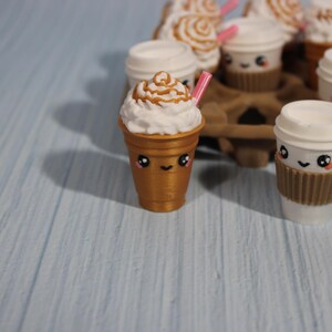 May include: A collection of miniature coffee cups with cute faces. Some cups have whipped cream and caramel drizzle, others have lids and brown sleeves. A pink straw is in one cup. The cups are arranged in a brown cardboard carrier.