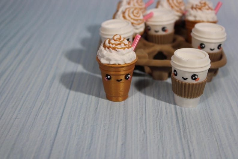 May include: A collection of miniature coffee cups with cute faces. Some cups have whipped cream and caramel drizzle, others have lids and brown sleeves. A pink straw is in one cup. The cups are arranged in a brown cardboard carrier.
