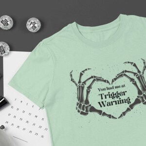 Könnte beinhalten: Ein hellgrünes T-Shirt mit einem schwarzen Skelett-Hand-Design, das eine Herzform bildet. Der Text "You had me at Trigger Warning" ist auf das Shirt gedruckt.