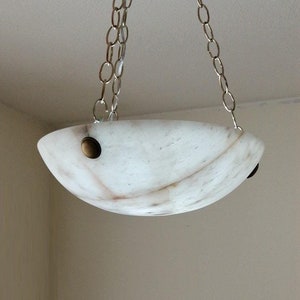 May include: A white alabaster pendant light fixture with a rounded, bowl-shaped shade. The fixture is suspended from the ceiling by a silver chain and has two brass accents on the sides.