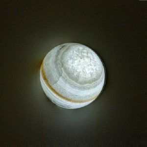 Op de afbeelding: Een witte en grijze geode bol met een lichtbruine band rond het midden. De geode wordt van binnenuit verlicht, waardoor een zachte gloed ontstaat.