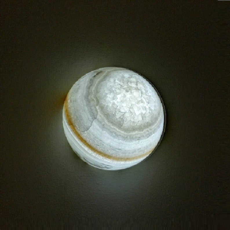 Op de afbeelding: Een witte en grijze geode bol met een lichtbruine band rond het midden. De geode wordt van binnenuit verlicht, waardoor een zachte gloed ontstaat.