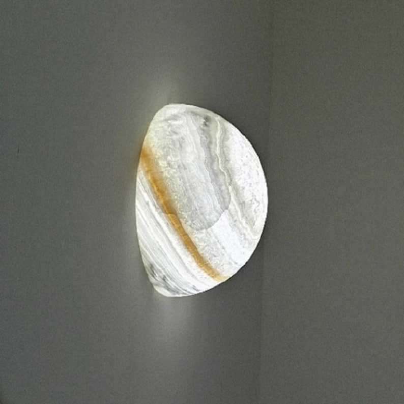 Op de afbeelding: Een witte en gouden albast wandlamp met een afgeronde vorm. De lamp wordt van binnenuit verlicht en geeft een warm licht.