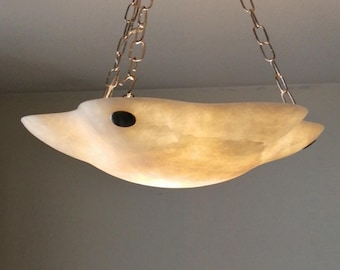 May include: A bowl-shaped pendant light fixture made of translucent, cream-colored stone with a smooth, organic edge. The light is suspended by three thin metal chains attached to the rim. A small, dark, circular accent is visible on the side.