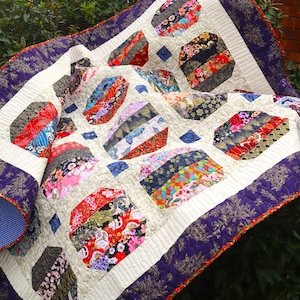 May include: A white quilt with a purple border and gold accents. The quilt is made up of various colourful fabric squares with geometric patterns. The quilt is folded and draped over a bush.
