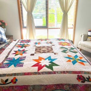 May include: A white quilt with a colourful patchwork design featuring stars and flowers. The quilt is on a bed with a white headboard and a window in the background.