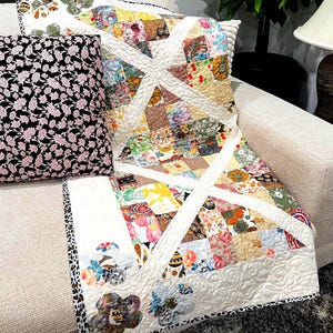 May include: A patchwork quilt with a white cross design and floral patterns in various colors. A black pillow with pink floral print rests beside the quilt on a beige sofa. The quilt's border has a black and white animal print.