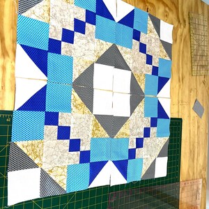 May include: A partially completed quilt top with a geometric design. The quilt features squares, triangles, and rectangles in shades of blue, white, gray, and gold. The quilt is on a green cutting mat with rulers.