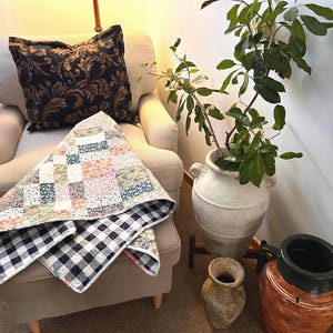 May include: A cozy living room scene featuring a beige armchair with a decorative pillow and a patchwork quilt. The quilt has a black and white checkered pattern on one side. A large white vase with greenery and other decorative pottery are also visible.