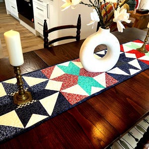 May include: A quilted table runner with a star pattern in shades of blue, white, red, and teal. The runner is on a wooden table with a vase of white flowers and two candlesticks.