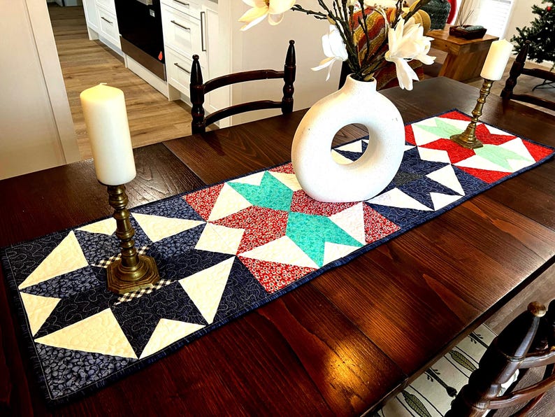 May include: A quilted table runner with a star pattern in shades of blue, white, red, and teal. The runner is on a wooden table with a vase of white flowers and two candlesticks.