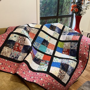 May include: A patchwork quilt with a black and white border and a pink floral pattern on the back. The quilt is made up of squares of different colors and patterns, including blue, green, red, and white.