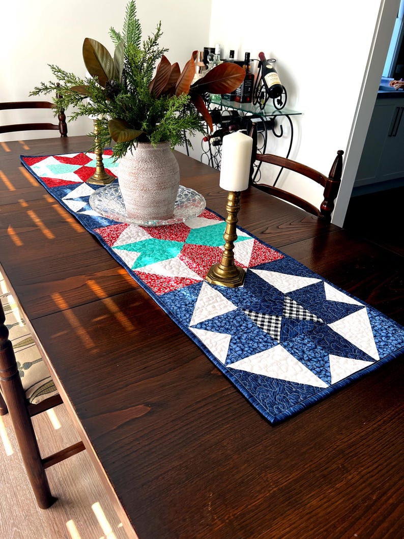 May include: A quilted table runner with a red, white, and blue star pattern. The runner is on a dark wood table with two candlesticks and a vase of greenery.