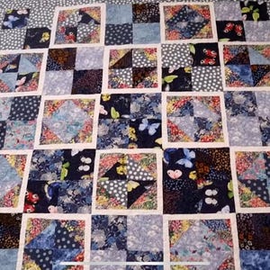 K&ouml;nnte beinhalten: Eine blaue und wei&szlig;e Patchwork-Decke mit einem Schmetterlingsmuster. Die Decke besteht aus Quadraten aus verschiedenen Stoffen, die jeweils ein anderes Design haben. Die Decke ist mit einem violetten Rand eingefasst.