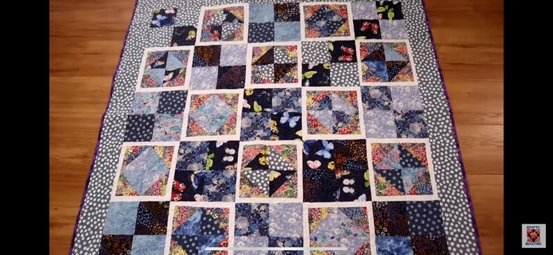 K&ouml;nnte beinhalten: Eine blaue und wei&szlig;e Patchwork-Decke mit einem Schmetterlingsmuster. Die Decke besteht aus Quadraten aus verschiedenen Stoffen, die jeweils ein anderes Design haben. Die Decke ist mit einem violetten Rand eingefasst.