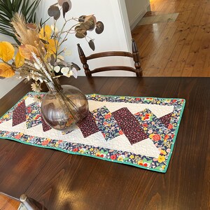 Op de afbeelding: Een donkerbruine houten tafel met een witte tafelrunner met bloemenpatroon. De tafelrunner heeft een groene rand en een chevron-patroon. Een heldere glazen vaas met gedroogde bloemen staat op de tafel.