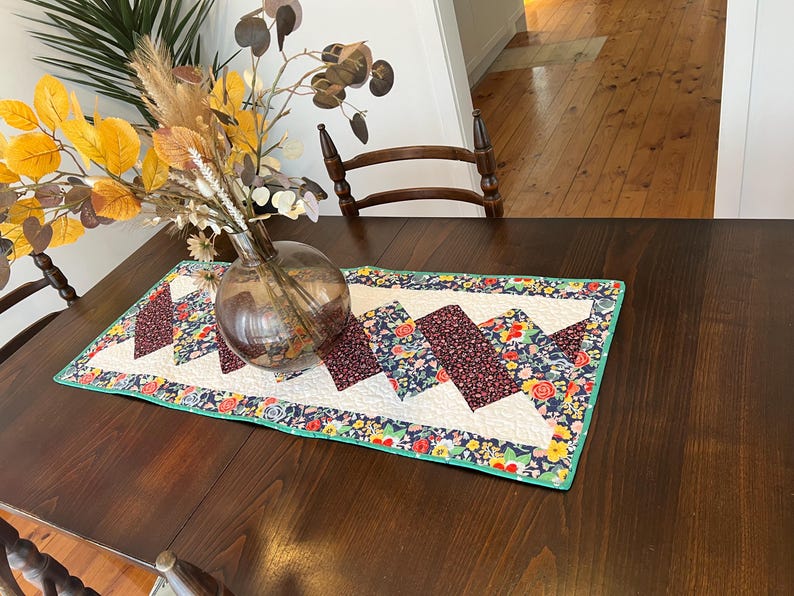 Op de afbeelding: Een donkerbruine houten tafel met een witte tafelrunner met bloemenpatroon. De tafelrunner heeft een groene rand en een chevron-patroon. Een heldere glazen vaas met gedroogde bloemen staat op de tafel.