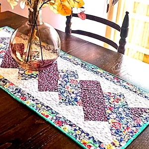 May include: A quilted table runner with a chevron pattern in shades of purple, teal, and white. The runner has a floral border and is displayed on a wooden table.
