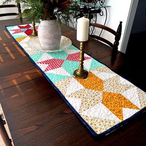 Op de afbeelding: Een gestikte tafelrunner met een geometrisch patroon in tinten rood, wit, geel, blauw en groen. De runner ligt op een donkerhouten tafel met een vaas met groen en kaarsen.