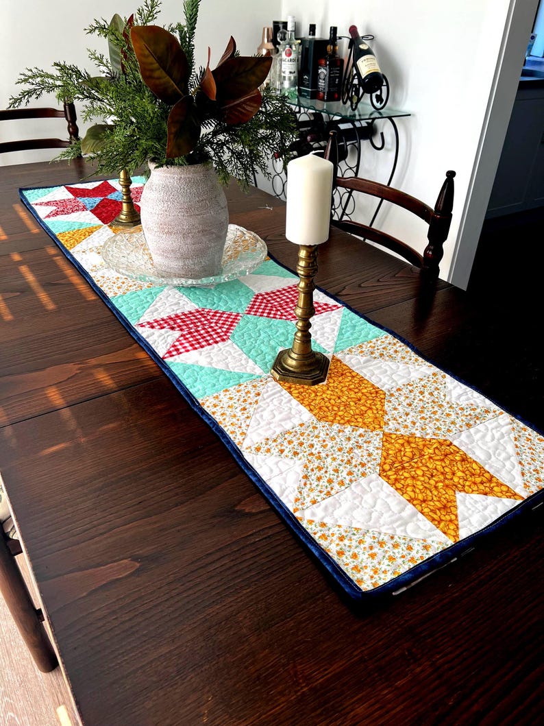 May include: A quilted table runner with a geometric pattern in shades of red, white, yellow, blue, and green. The runner is on a dark wood table with a vase of greenery and candles.