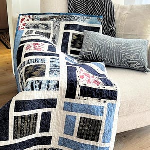 Op de afbeelding: Een handgemaakte quilt met een geometrisch ontwerp in tinten blauw, wit en zwart is over een beige fauteuil gedrapeerd. De quilt heeft rechthoekige en vierkante patronen. Een decoratief kussen met een grijs patroon en een zwart-wit patroon kussen liggen ook op de stoel.