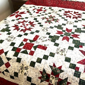 May include: A handmade quilt with a festive design. The quilt features a pattern of red and green stars and squares on a white background. The border is a solid red fabric. The quilt is likely made of cotton.
