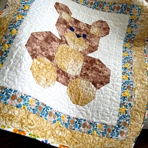 May include: A handmade quilt featuring a brown teddy bear design on a white background. The bear is made of various brown and tan patterned fabrics. The quilt has a floral border with blue, yellow, and white flowers, and measures approximately 40 x 40 inches.
