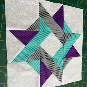 May include: A square quilt block featuring a star design. The star is composed of teal, purple, and gray polka-dot fabric pieces, set against a white floral background. The block is on a green cutting mat.