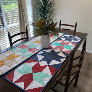 May include: Three colorful quilted table runners with geometric star patterns in shades of red, blue, green, yellow, and white. The runners are laid out on a wooden table with two chairs.