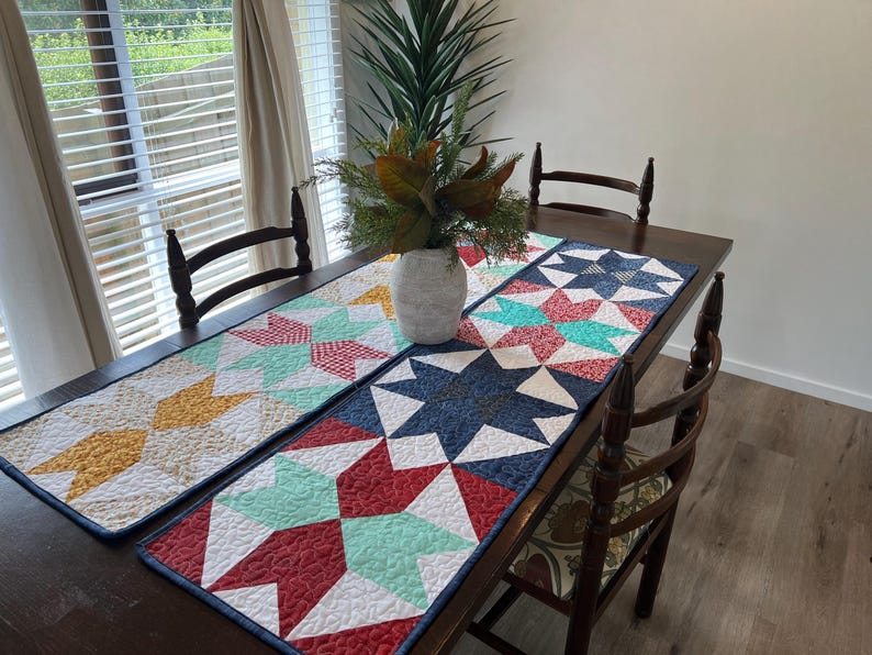 May include: Three colorful quilted table runners with geometric star patterns in shades of red, blue, green, yellow, and white. The runners are laid out on a wooden table with two chairs.