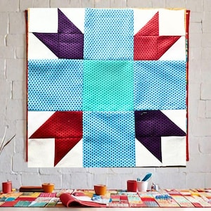 May include: A colourful quilt block with a star design. The block is made of red, blue, green, purple and white fabric. The fabric has a polka dot pattern.