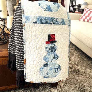 May include: A quilted snowman-themed decorative cover with a white background and blue floral accents. The snowman has a red and black top hat. The cover is likely for a chair or ottoman, adding a festive touch to home decor.