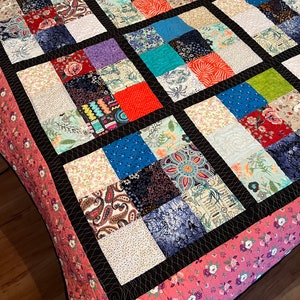 May include: A patchwork quilt with a black border and a pink floral pattern on the back. The quilt is made up of squares of different fabrics in various colours and patterns.