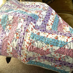Op de afbeelding: Een kleurrijke quilt met een bloemenpatroon in roze, paarse, blauwe en witte tinten. De quilt heeft een diagonaal ontwerp met rijen stof die aan elkaar zijn genaaid. De quilt is waarschijnlijk gemaakt van katoen of een vergelijkbare stof.