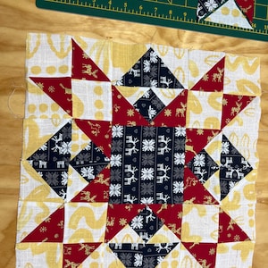 May include: A patchwork quilt block in progress, featuring a central star design. The block incorporates red, navy, and white fabrics with reindeer and snowflake patterns. Yellow and white patterned fabric forms the background. Additional cut pieces are visible.