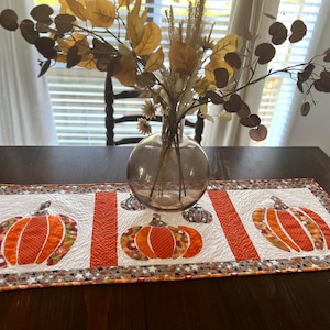 May include: A white table runner with three orange and brown pumpkin appliques. The runner is on a dark wood table with a vase of dried leaves in the background.