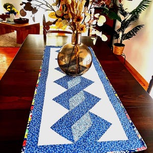Op de afbeelding: Een blauwe en witte gestikte tafelrunner met een ruitpatroon. De runner heeft een bloemige rand en wordt getoond op een houten tafel met een vaas met gedroogde bloemen.