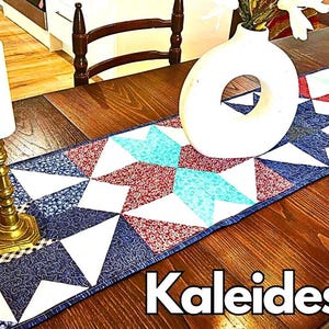 May include: A table runner with a kaleidoscope pattern in shades of blue, red, and white. The runner is made of fabric and has a quilted design. The runner is on a wooden table with a vase and candles.