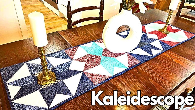 May include: A table runner with a kaleidoscope pattern in shades of blue, red, and white. The runner is made of fabric and has a quilted design. The runner is on a wooden table with a vase and candles.