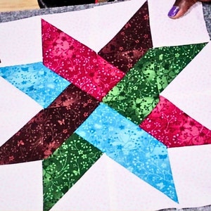 May include: A patchwork quilt block featuring a star design with six points. The star is made of six different fabric pieces in shades of red, green, brown, and blue. The fabric has a floral pattern.