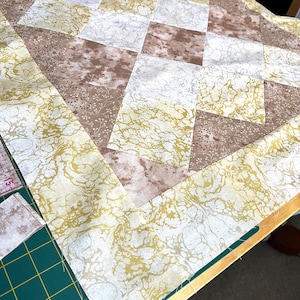 May include: A patchwork quilt in progress, with a geometric design of squares and triangles. The quilt uses fabrics in shades of brown, cream, and gold, with a marbled or floral design. The quilt is on a green cutting mat.