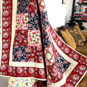 May include: A red, white and blue floral quilt with a patchwork design. The quilt is folded and draped over a sofa.