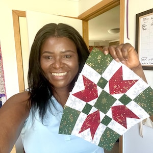 May include: A smiling person holds a Christmas-themed quilt block. The block features red star shapes with the word "joy" and "hope" on them, set against a green and white background with snowflake and leaf patterns. The block is a square shape.