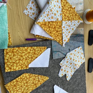 May include: A close-up of fabric scraps in various patterns and colors, including white, yellow, and orange. The fabric is cut into different shapes and sizes, and some of the pieces are layered on top of each other. The fabric is arranged on a gray surface, and there are tools for sewing nearby.