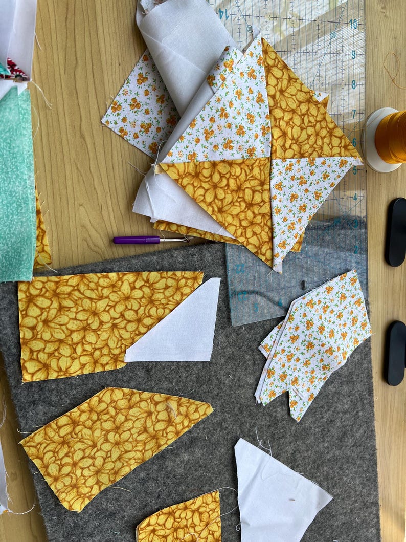May include: A close-up of fabric scraps in various patterns and colors, including white, yellow, and orange. The fabric is cut into different shapes and sizes, and some of the pieces are layered on top of each other. The fabric is arranged on a gray surface, and there are tools for sewing nearby.